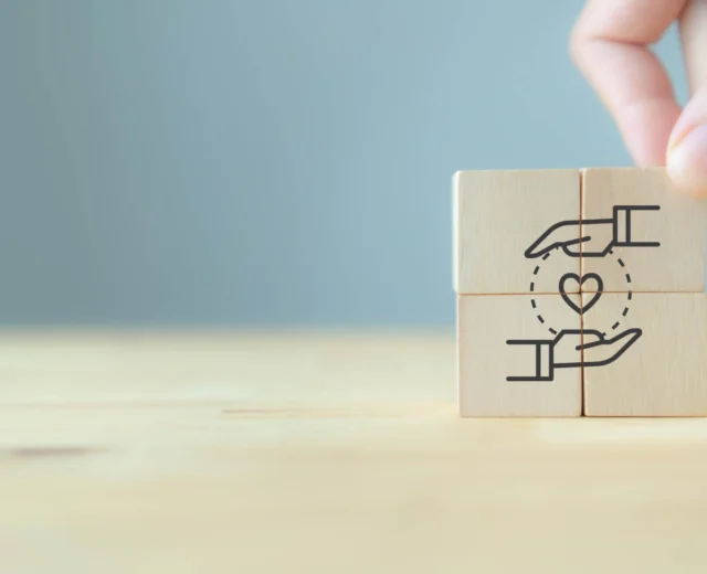Four wooden blocks with a hand, heart, and hand design are arranged to form a complete image. A hand is placing the final block.
