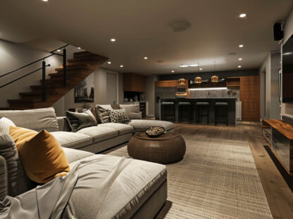 A modern living room with a sectional sofa, cushions, a large TV, and a pouf. The adjacent kitchen area has bar stools and pendant lights. A staircase is visible on the left.