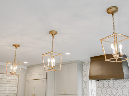 Three gold geometric pendant lights hang from the ceiling in a modern kitchen with white cabinets and a geometric backsplash.