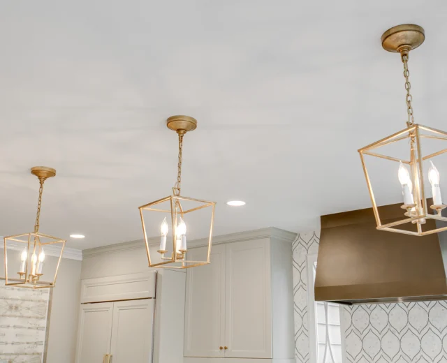 Three gold geometric pendant lights hang from the ceiling in a modern kitchen with white cabinets and a geometric backsplash.