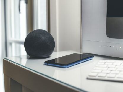 A smart speaker, smartphone, and keyboard are placed on a glass desk near a computer monitor.