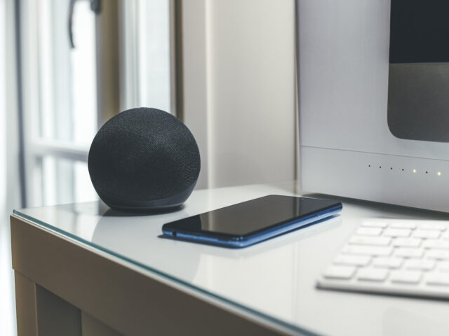 A smart speaker, smartphone, and keyboard are placed on a glass desk near a computer monitor.