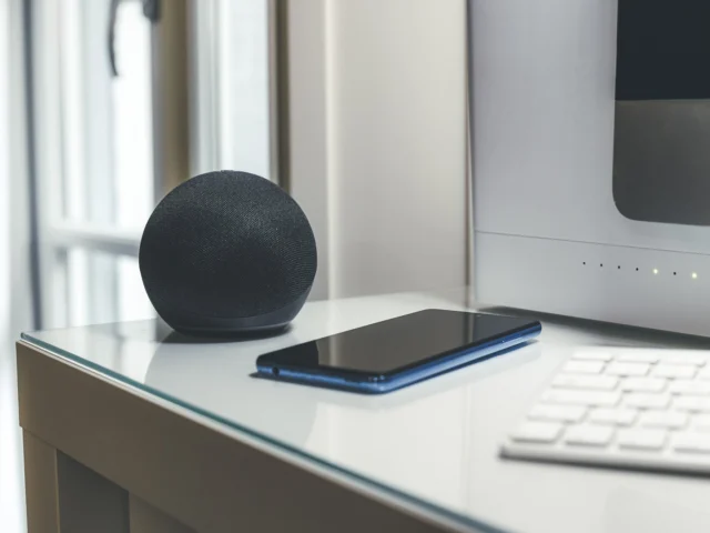 A smart speaker, smartphone, and keyboard are placed on a glass desk near a computer monitor.