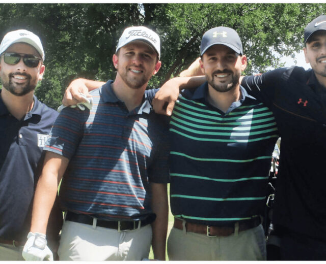 Four men in golf attire stand together on a golf course, smiling at the camera.