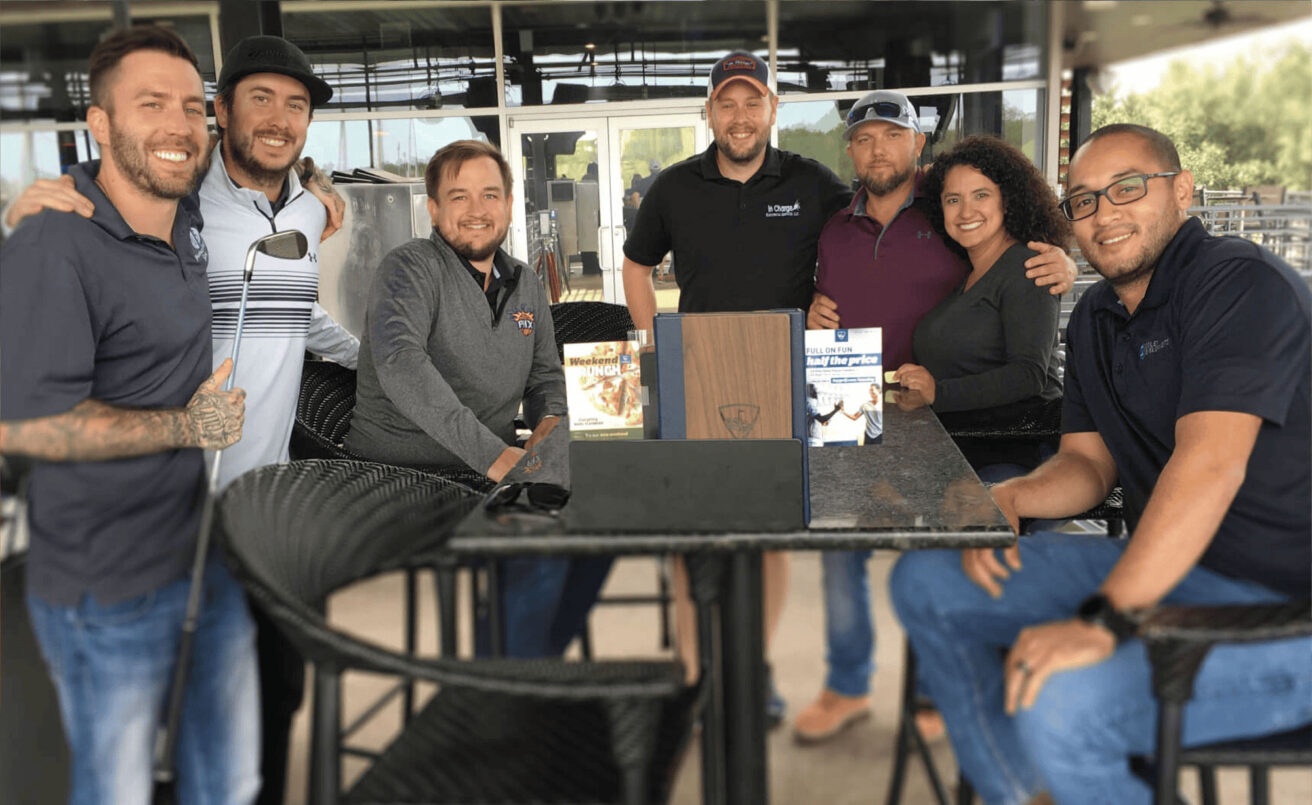 A group of seven people posing together around a high table at an outdoor patio, smiling at the camera.