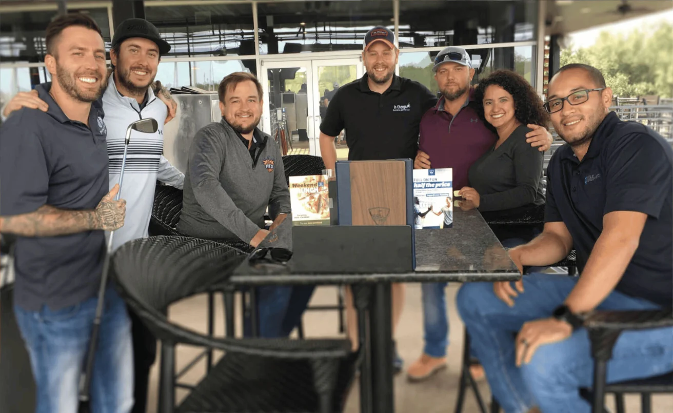 A group of seven people posing together around a high table at an outdoor patio, smiling at the camera.