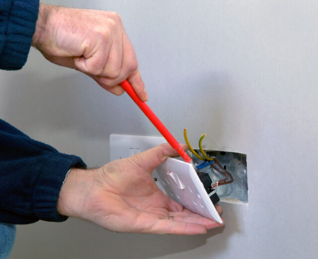 Person using a screwdriver to install an electrical outlet into a wall with exposed wiring.
