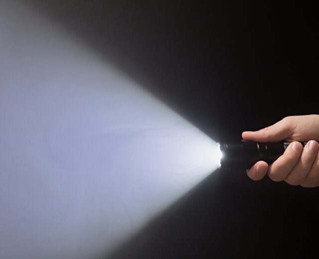 Black flashlight with a beam of light in male's hand isolated from right side of the frame on black background.