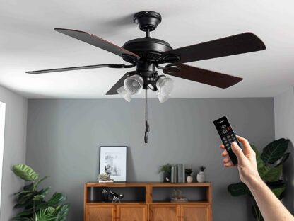 A hand holding a remote points at a black ceiling fan with four blades and lights||mounted in a modern room with gray walls and plants.