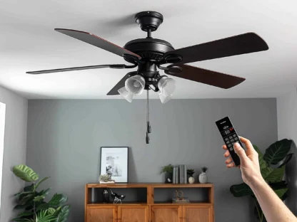 A hand holding a remote points at a black ceiling fan with four blades and lights||mounted in a modern room with gray walls and plants.
