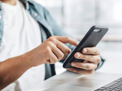 Person using a smartphone while seated at a desk with a laptop.||