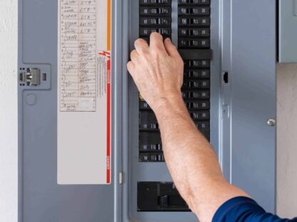 A person adjusts a breaker switch in an open electrical panel.|Electrical panel with wires in a wooden framework of a house under construction.|