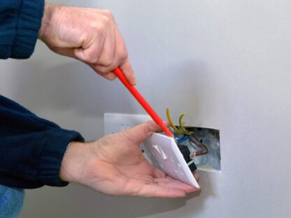 Person using a red screwdriver to install or repair a wall electrical outlet||with exposed wires visible inside the wall cutout.