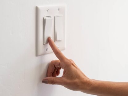 A hand reaching to flip a light switch on a white wall.||