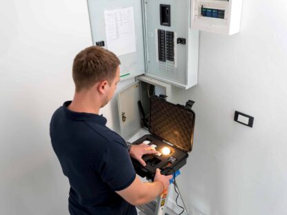 A man stands on a ladder inspecting an electrical panel with a testing device in a case|