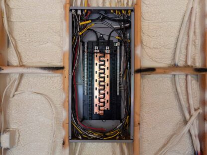An open electrical panel with visible wiring and circuit breakers is set against a wall with insulation foam.||