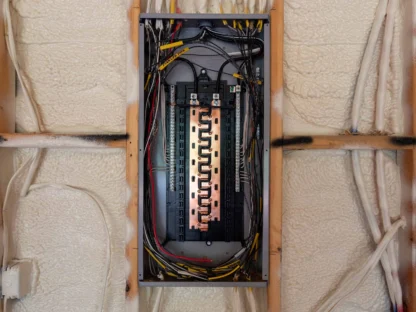 An open electrical panel with visible wiring and circuit breakers is set against a wall with insulation foam.||